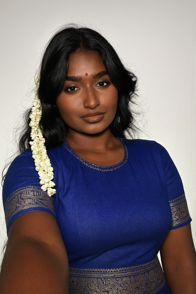 A woman with long, dark, wavy hair, adorned with a string of white flowers on her left side, looks directly at the viewer. She is wearing a royal blue top with a round neckline and short sleeves, featuring gold patterned borders around the neck, sleeves, and hem. A small red bindi is on her forehead. Her expression is calm and direct. The background is a plain, light-colored wall.