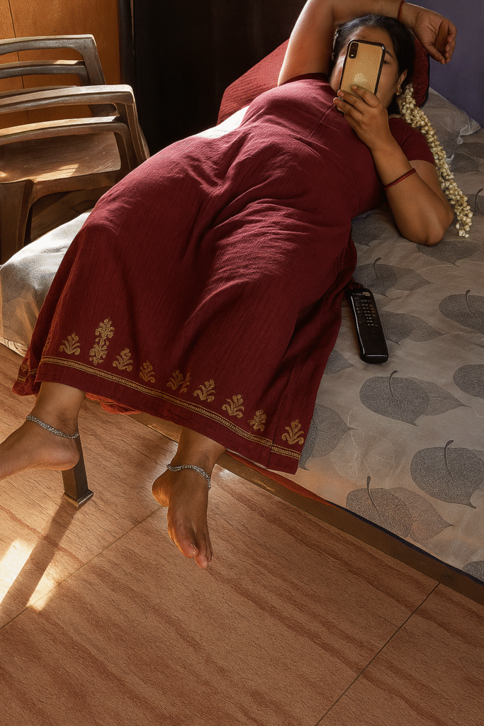 A full-body shot captures a woman lying on her side on a bed, looking at her phone. She is wearing a dark red dress or nightgown with gold floral embroidery along the hem. Her right arm is bent and resting on the pillow, while her left arm is holding a light-colored smartphone in front of her face. A string of white flowers is visible in her hair, resting on the pillow. Her bare feet, adorned with silver anklets, are visible at the bottom of the image, extending onto a light brown wooden floor, part of which is bathed in sunlight. A dark remote control lies on the patterned bedspread near her. In the background, a wooden chair is partially visible. The lighting in the room is a mix of bright sunlight and softer indoor illumination.