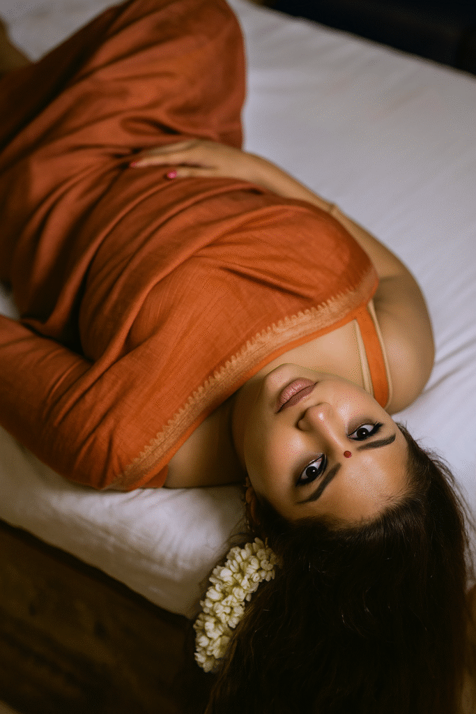 An overhead shot of a woman with dark hair and medium brown skin, lying on a white surface and looking up at the camera with a soft expression. A string of white jasmine flowers is tucked into her hair behind her head on the right side. She is wearing an orange-brown saree with a subtle pattern and a light-colored border, draped loosely around her. A small red bindi is on her forehead, and her eyes are framed with dark eyeliner. Her left hand is resting gently on her stomach. The background is a plain white surface, likely a bed.