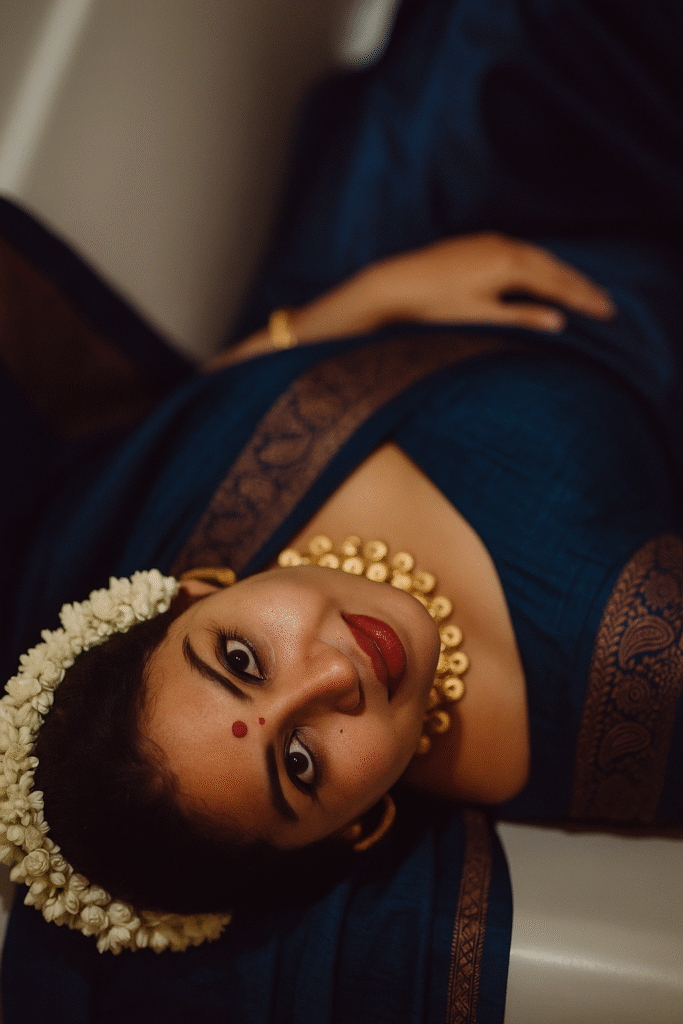 An overhead shot of a woman with dark hair and medium brown skin, lying down and looking up at the camera with a slight smile. A string of white jasmine flowers frames her head on the left side. She is wearing a deep teal-blue saree with a gold patterned border, and a prominent gold beaded necklace is around her neck. A small red bindi is on her forehead, and her lips are colored with a bold red lipstick. Her eyes are wide and framed with dark eyeliner. Her right hand is visible in the upper part of the frame. The background is dark and indistinct, suggesting an indoor setting.