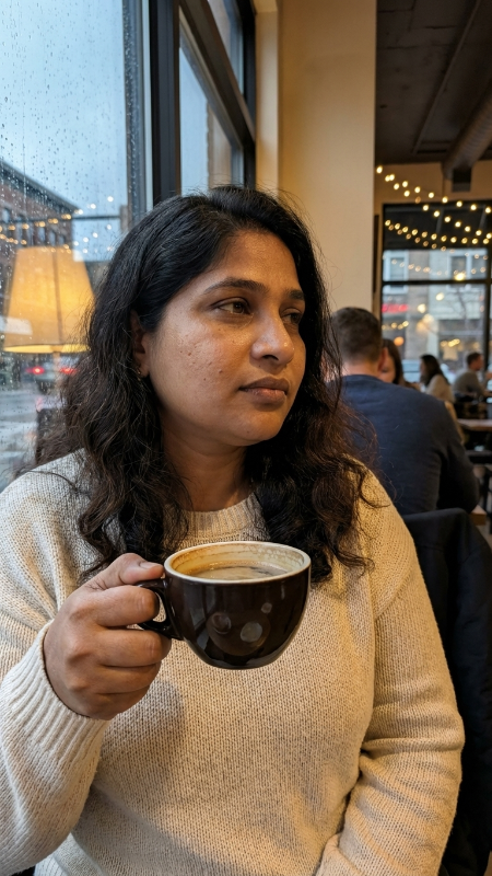 Create a realistic close-up photo of a modern young woman sitting inside a cozy coffee shop near a large window. She is wearing a simple cream-colored sweater and has natural makeup. She is holding a cup of coffee and looking outside thoughtfully. Add warm indoor lighting, blurred background lights, realistic skin details, natural hair texture, and soft rain visible outside the window.